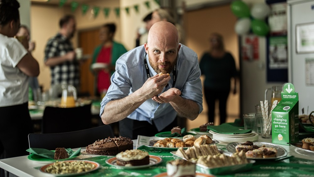 A man is leaning over a Coffee Morning table and taking a bite from a pastry. The man has on a blue shirt. The table has many different desserts and other treats on it. Behind the man are other people eating food and socialising.