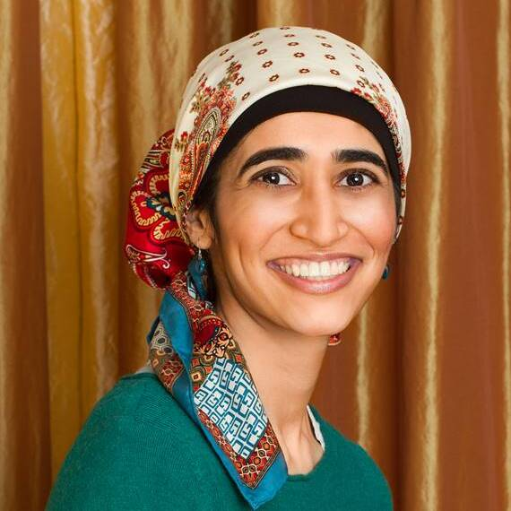 A headshot of Baraka Khan from the shoulders up. She is wearing a teal knitted top and a patterned head scarf. She is smiling. Behind her is a burnt orange curtain as a backdrop.