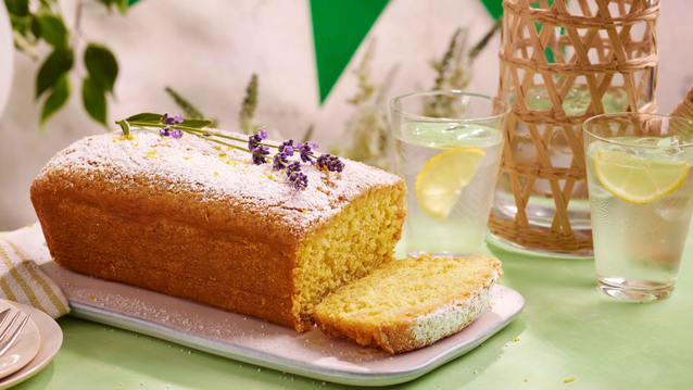 A golden lemon drizzle loaf cake dusted with powdered sugar and sprigs of fresh lavender, resting on a white rectangular plate. A slice has been cut to reveal its soft, fluffy interior.