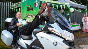 Steve is leaning on a motorbike seat with his legs on the handles. He is holding a teacup in one hand and plate with a cupcake in the other. He appears to be outside an an event. 