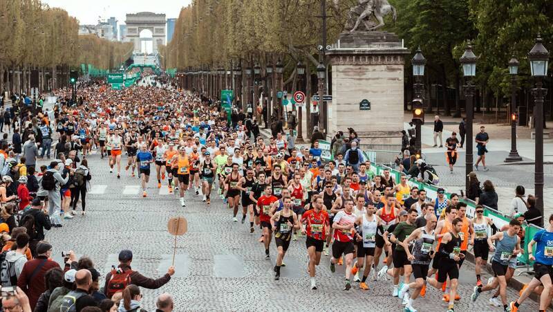 Many runners are running down a road away from the Arc de Triomphe. They are taking part in the Paris marathon.