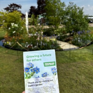 A person with red nail varnish on is holding up a leaflet for a Macmillan event. Behind the leaflet is a garden that is in a circle shape. It has been roped off. It has different plants in it and a gravel path.