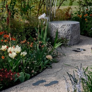 A set of fake footprints are on a gravel path in a garden. They lead to a stone chair. The garden has different plants and flowers.