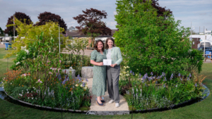 Kerianne Fitzpatrick (left) and TJ Kennedy (right) are standing on a path in the Macmillan Legacy Garden at the at RHS Malvern Spring Festival. They are both holding a piece of paper. The garden is round and had many plants.