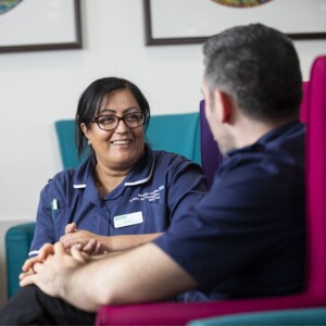 Suki, a Macmillan professional, is sitting in a chair at a Macmillan Centre. They are wearing a blue NHS uniform. In the chair opposite from them is a patient. They are talking to each other.