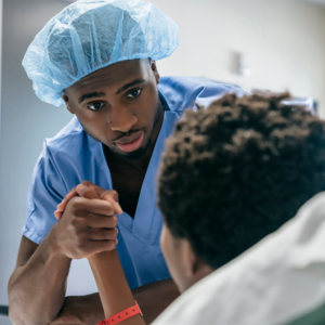 A nurse in blue scrubs and wearing a blue scrub cap is holding the hand of a patient who is laying down in a medical bed.