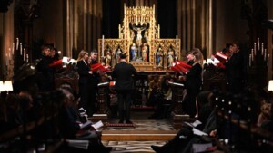 A chorus is singing inside a cathedral. They are standing on the left and right side of a conductor. The singers are holding red song booklets.