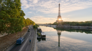 A view of the Eiffel tower near the Seine. A person is riding their bicycle on the bike path next to the river. There are boats parked along the river and vehicles driving on the other side of the cyclist.