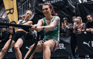 A person wearing a green Macmillan fitness top is pulling a black rope. They are leaning back as they pull the gym rope. They appear to be taking part in a fitness event indoors.