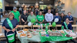 A group of Welcome Break employees are standing together next to a table decorated for a Coffee Morning. There are different foods on the table and one person is standing holding a Macmillan branded donation bucket.