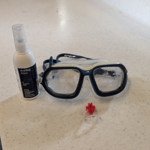 A pair of swimming googles on a counter. Next to them are defog googles and a swimming nose plug.