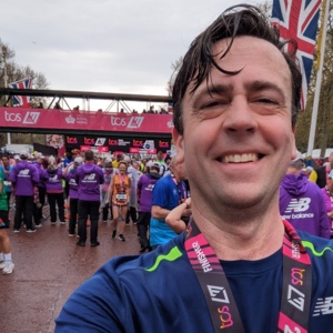 A selfie of Nick. He is smiling. Nick has finished running a marathon. He has a blue running top on and a finisher's medal on a lanyard. He is outside.