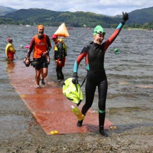 Heather is stepping out of lake onto a rocky beach. She has one arm in the arm and the other is holding a flotation device. Heather is wearing a wet suit, a swimming cap and googles.