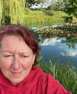 A selfie of Gill. She is outside in front of a lake. She has short hair and is wearing a red hoodie.