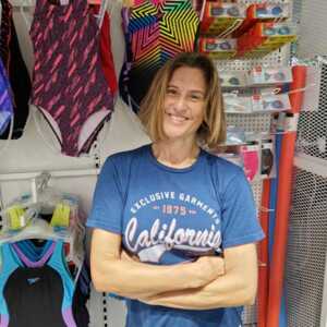 Johanna has short brown hair. She is standing up with her arms across her stomach. She is smiling and is wearing a blue top and pink trousers. She is standing in front of different swimming equipment including swimming costumes and goggles.
