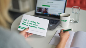 A person is holding a Macmillan Free Wills Service guide in their left hand. In the right hand they are holding a pen over an open notebook that is on a table. In the background is a laptop, a cup of coffee and a glass of water.