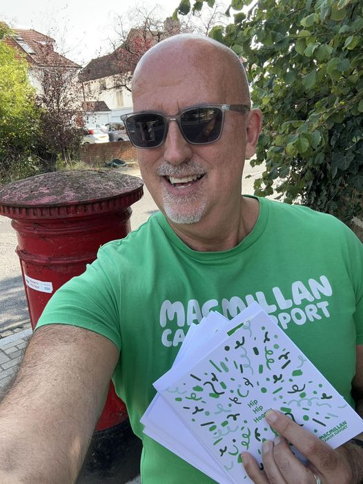 A Macmillan volunteer is taking a selfie. They are holding a thank you card in their hands and standing in front of a red post box. They are wearing a green Macmillan branded top and sunglasses.