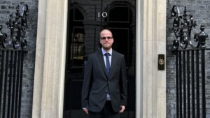 Nathaniel is standing in front of 10 Downing Street, the official residence of the Prime Minister. He is wearing a black suit with a dark blue tie. 