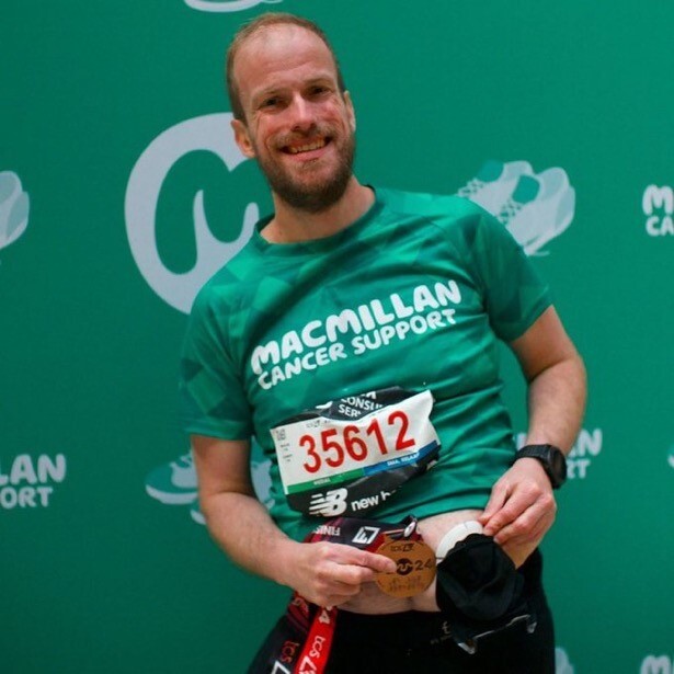 Nathan is wearing a green Macmillan branded fundraising top. He is smiling and holding a marathon medal. You can see Nathaniel's colostomy (stoma) bag. He is standing in front of a Macmillan branded background.