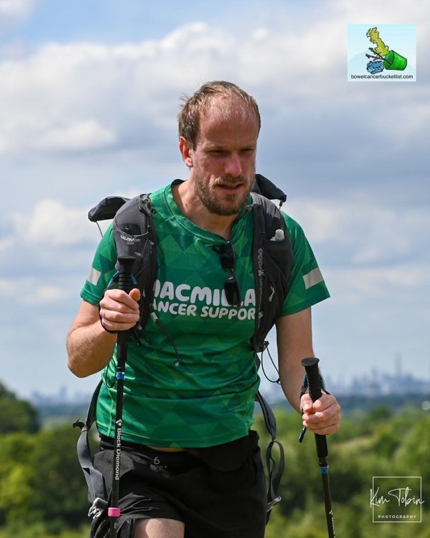 Nathanial is holding walking sticks and is hiking. He has on a green Macmillan branded top and dark shorts. In the background is trees and a city skyline.
