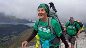 Two people are hiking along a rocky path. They are both wearing green Macmillan branded tops. They are also wearing other hiking gear like backpacks.