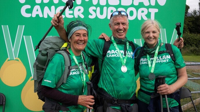 A group of three people are posing for a photo. They are wearing hiking gear including green Macmillan branded tops and rucksacks. One hiker has their arms in the air and they are holding walking sticks.