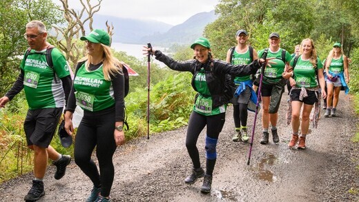 A group of hikers are walking along a muddy path. They are wearing green Macmillan tops and other hiking gear. One hiker is holding walking sticks.