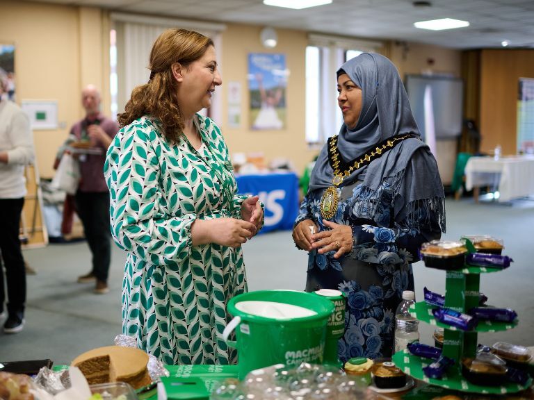 Two women in conversation, one has brown hair and is wearing a green patterned dress. The other is wearing a blue hijab and floral dress, she's also wearing a ceremonial gold chain around her neck