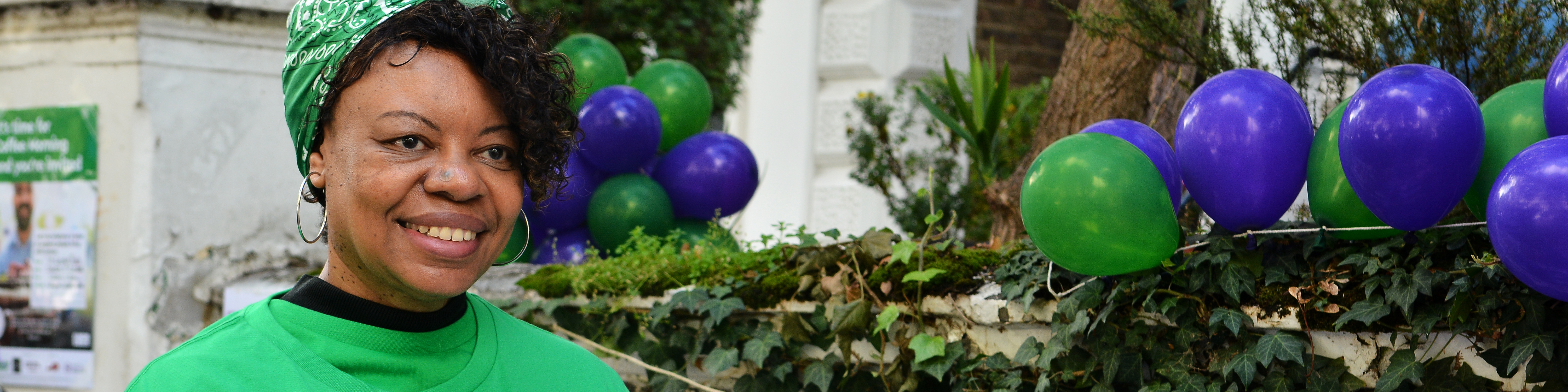 Debbie is wearing a green Macmillan branded Coffee morning t-shirt. She has hair hair in a green bandana too. She is standing outside holding a paper coffee cup. Behind her is a table decorated for a Coffee Morning event. 