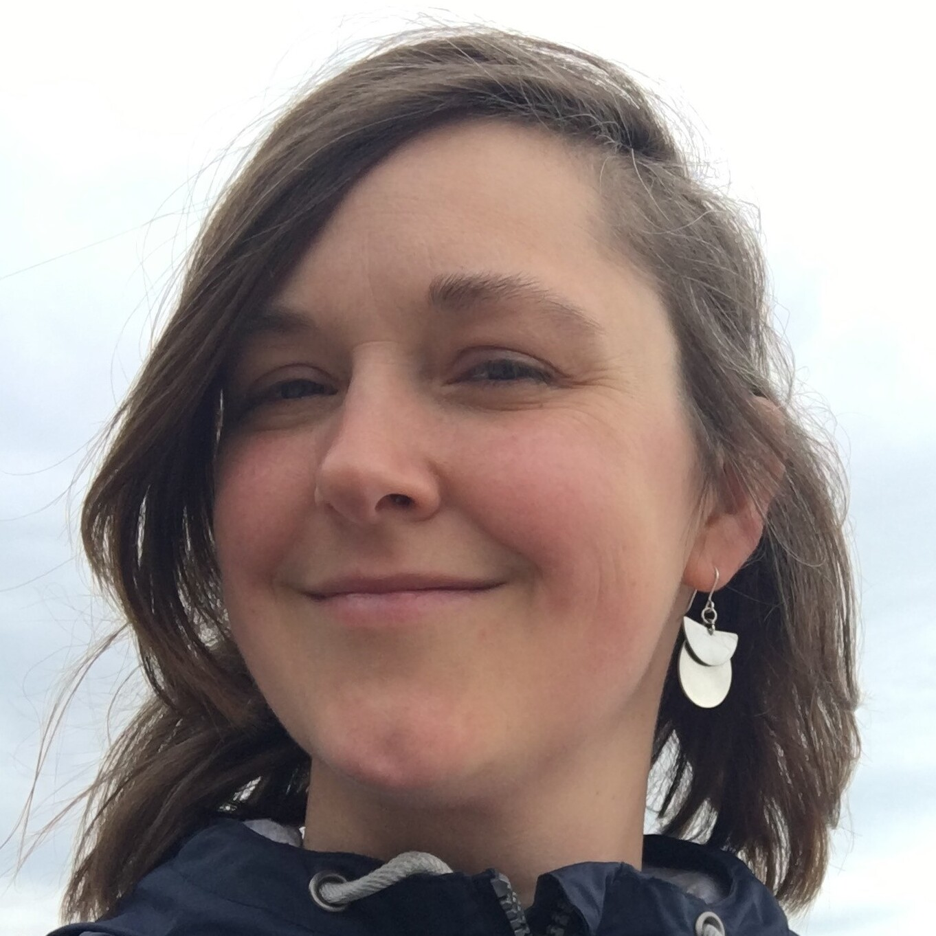 Martha has brown hair and wears a blue waterproof jacket. She is standing on a beach smiling.