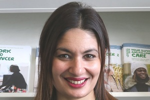 Azmina, a Macmillan Recovery Package Project Manager, smiling in front of a shelf of Macmillan booklets.