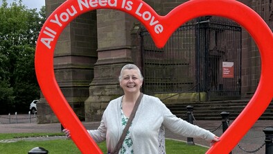 Stephanie is standing outside behind a large heart shaped sculpture. She is wearing a grey cardigan, a patterned top and pink trousers. The sculpture has the words All You Need is Love written on them.