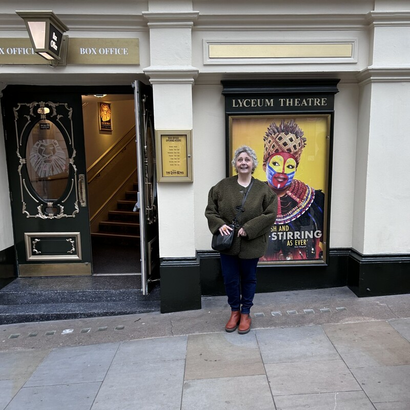 Stephanie is standing outside the Lyceum Theatre. She is wearing a long, dark green jacket, blue trousers and brown shoes. She has short, grey hair.