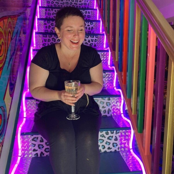 A woman is sitting on stairs with neon lights running up the sides. She's laughing and holding a glass of wine.