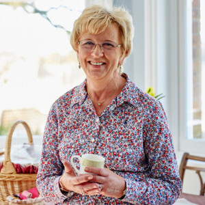 Woman with short blonde hair and glasses stands in a bright kitchen holding a coffee mug.