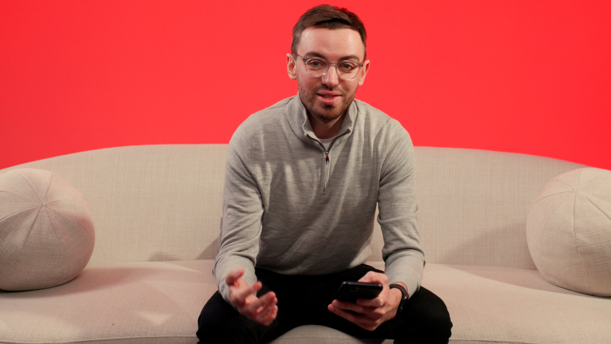 A man in his 30s sat on a sofa with dark hair, glasses and beard.