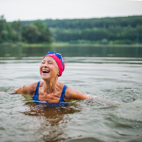 An older person is swimming in a lake. They are wearing a pink swimming cap, blue swimming goggles and a blue swim suit.