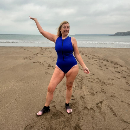 Lesley is standing on a sandy beach. She is wearing a dark blue swim suit and swim boots. She has one arm in the air. Behind her is the sea.