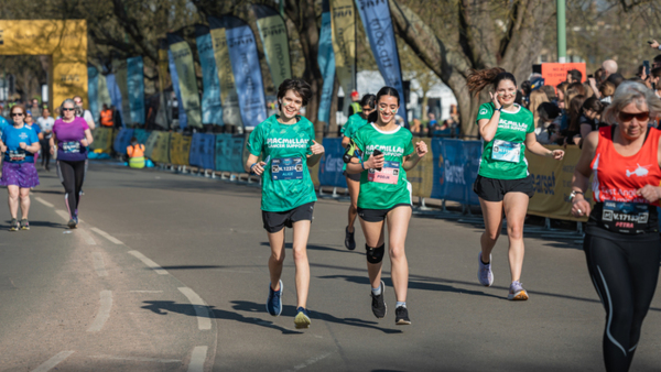 A group of runners are taking part in a race. Some of them are wearing green Macmillan branded sports tops. There are people cheering on either side of the running course.