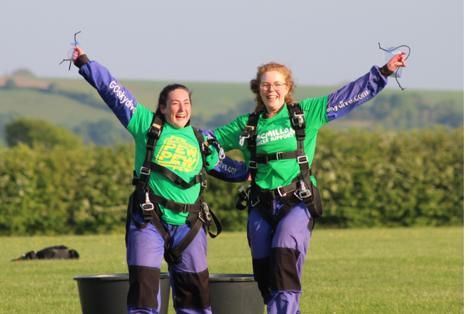 Two women in green Macmillan Cancer Support shirts and blue skydiving suits celebrate after landing smiling with arms raised