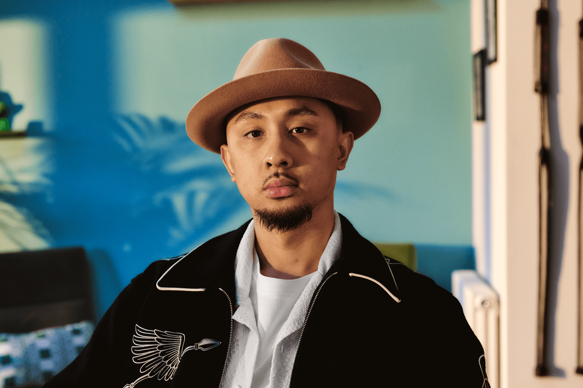 A young man of Asian heritage is sitting at home looking directly to camera. He has a goatee beard and is wearing a hat.