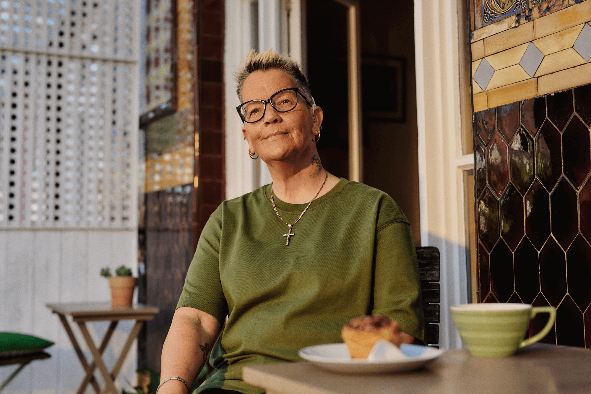 A woman is sitting at a patio table with a coffee and cake. She is wearing glasses and a hearing aid.