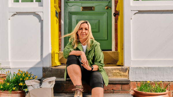 A woman in is sitting on a front door step looking at the camera. She is wearing hiking boots and has a backpack