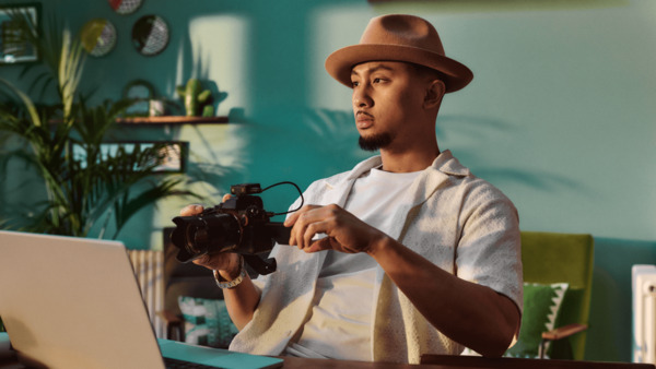 A young man of Asian heritage is sitting at home at a desk in front of a laptop. He's holding a camera. There are plants and ornaments in the background.