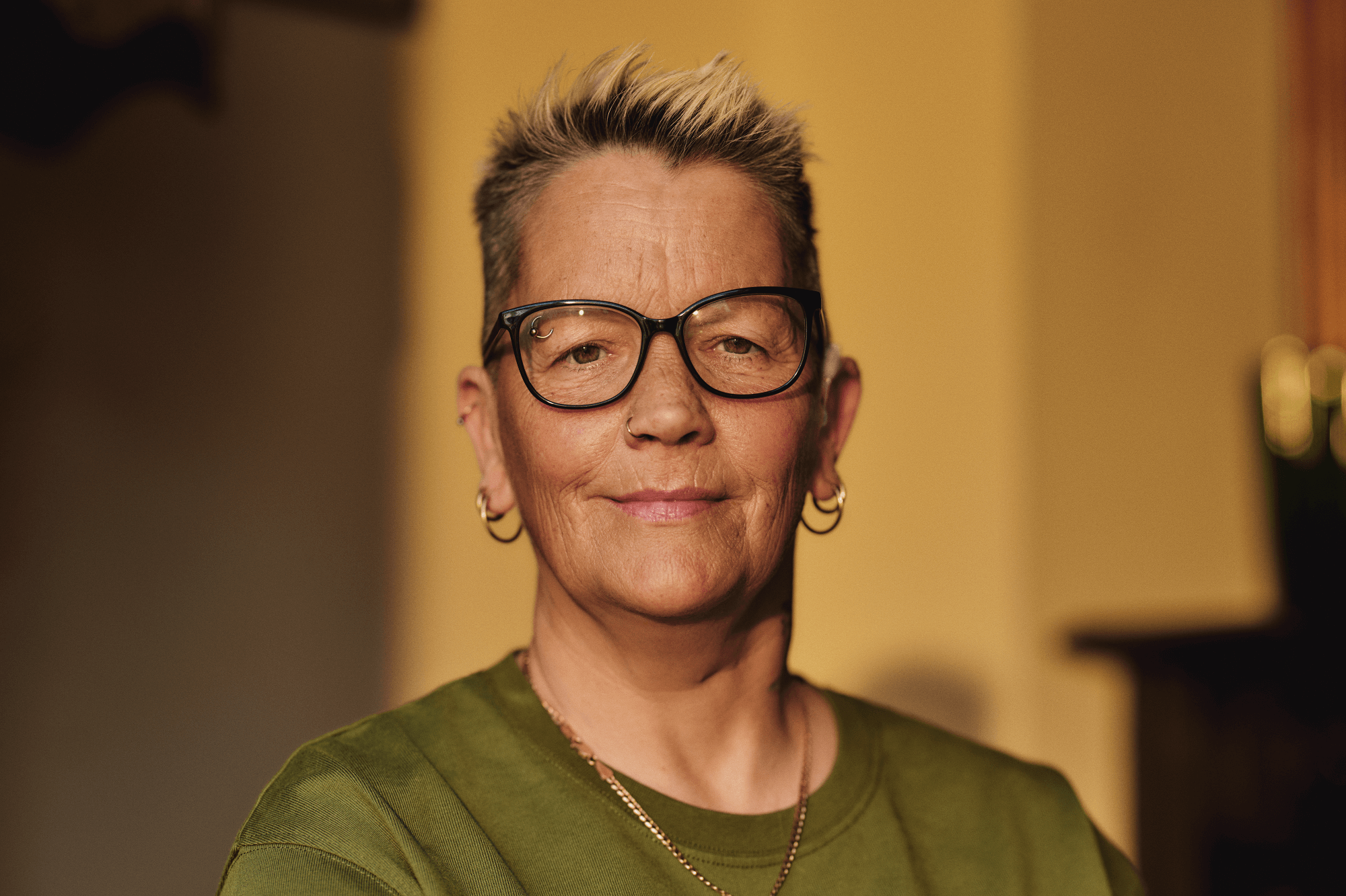 A close-up of a woman looking to camera.  She has short hair and is wearing glasses and has piercings in her ears, nose and eyebrow. Her hearing aid is just visible.