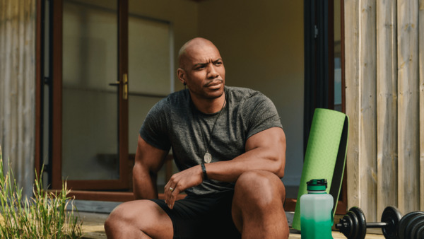 A Black man wearing sports clothes is sitting outside on some decking, He is a body builder and has weights and a water bottle beside him.