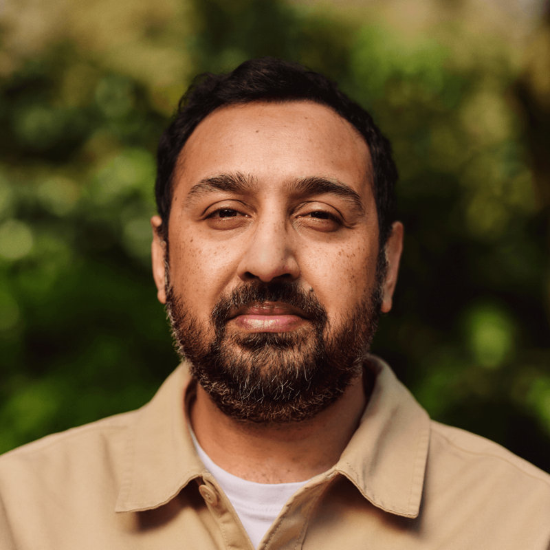 A man is outside looking at the camera. He's wearing a light-coloured shirt and has a beard. There are trees behind him.