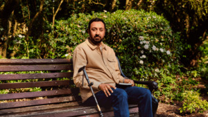 A man is sitting on a park bench. He has a walking stick next to him and a book in his hand.