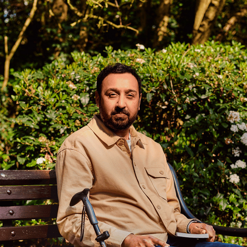 Mohammad sitting on a park bench. He has a walking stick next to him and a book in his hand.