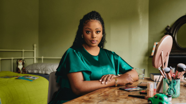 A young Black woman is sitting at home at a table which has make up on. There is a bed in the background with children's toys and books on.